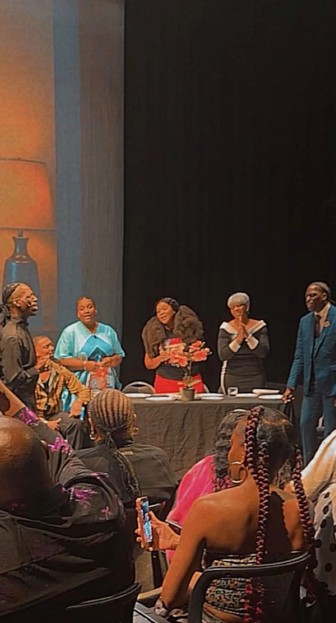 Cast members perform around a table onstage as a seated audience watches