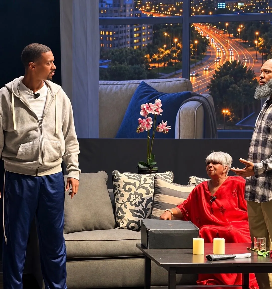 Three actors perform in a dramatic living-room stage scene, with one standing, one seated in red, and another gesturing